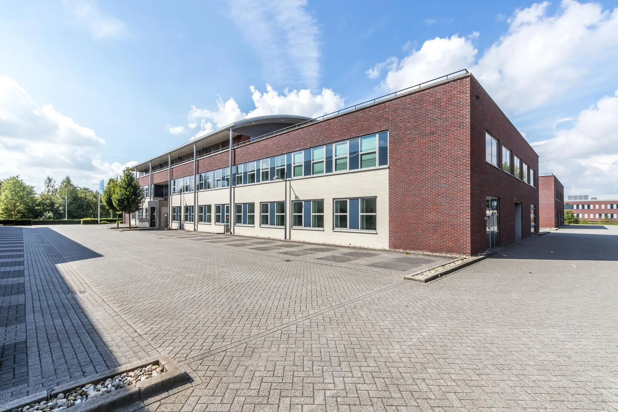 Modern bedrijfsgebouw De Wel met roodbruine en witte gevels op een ruim, bestraat terrein onder een blauwe lucht.